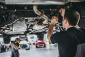 Two handsome mechanics in uniform are working in auto service with lifted vehicle. Car repair and maintenance.